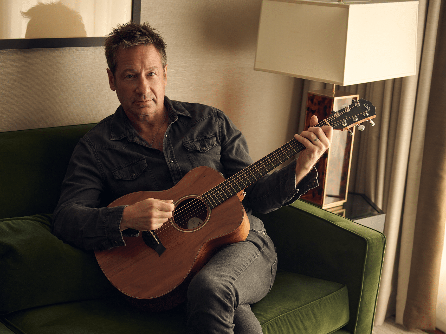 David Duchovny holding an acoustic guitar, sitting on a green sofa